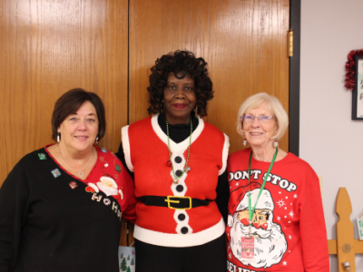 Three women smile together during the Worth Township Senior Christmas Party, wearing festive holiday sweaters and standing in front of Christmas decorations.